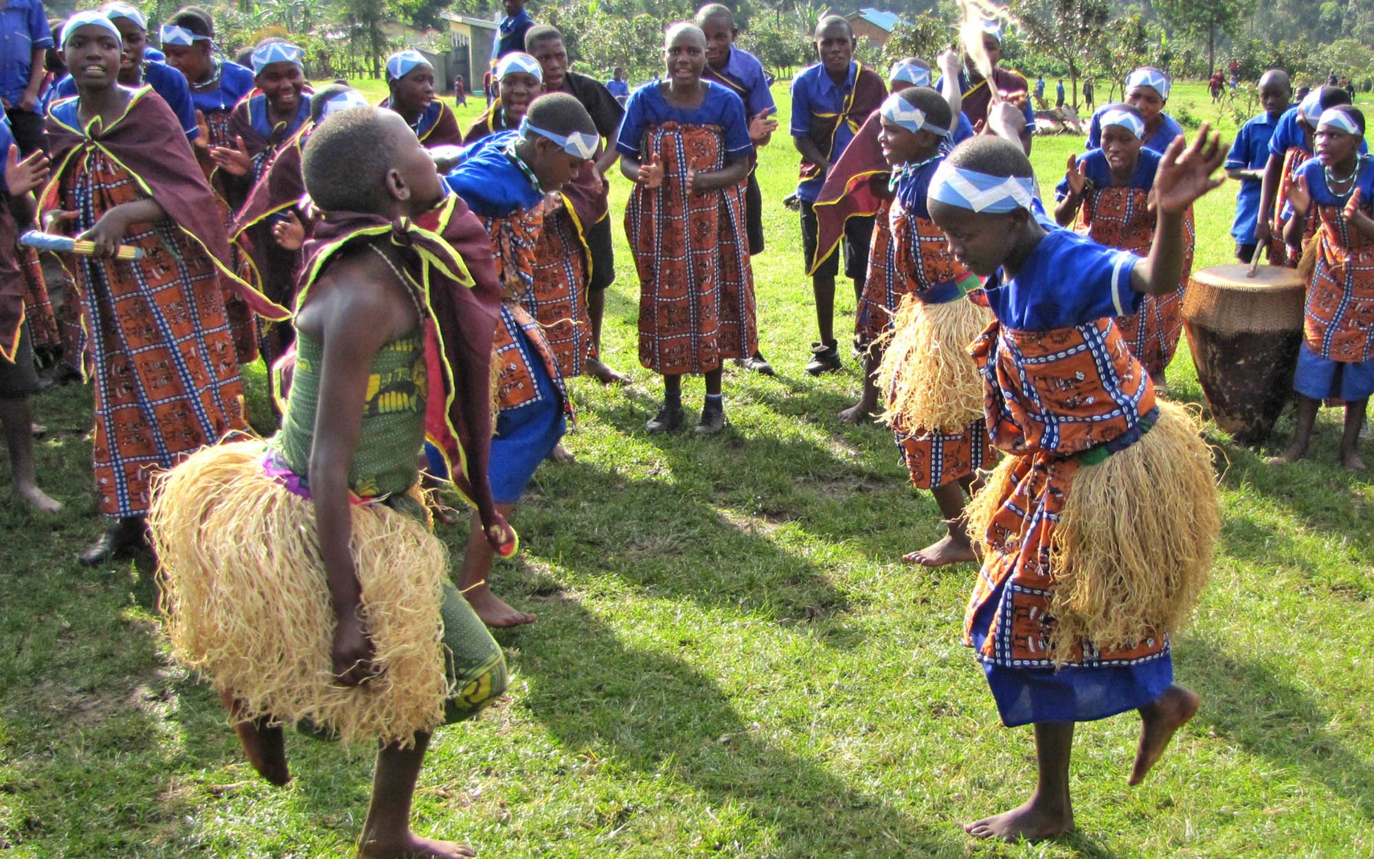 Children wearing colorful blue blue tunics and red scores with grass skirts engage in a traditional dance in Rubuguri Uganda