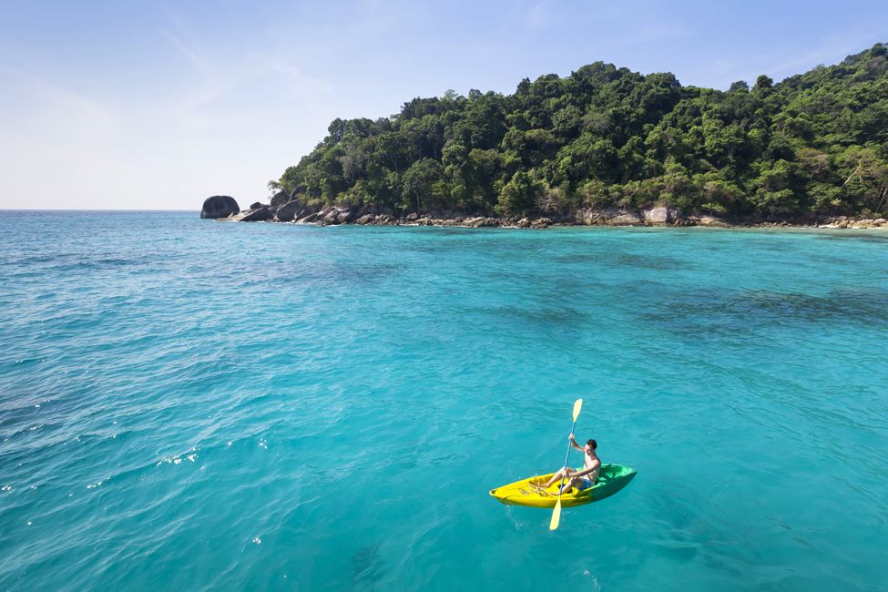 Young adult paddles yellow kayak in turquoise waters with tree-covered island in background