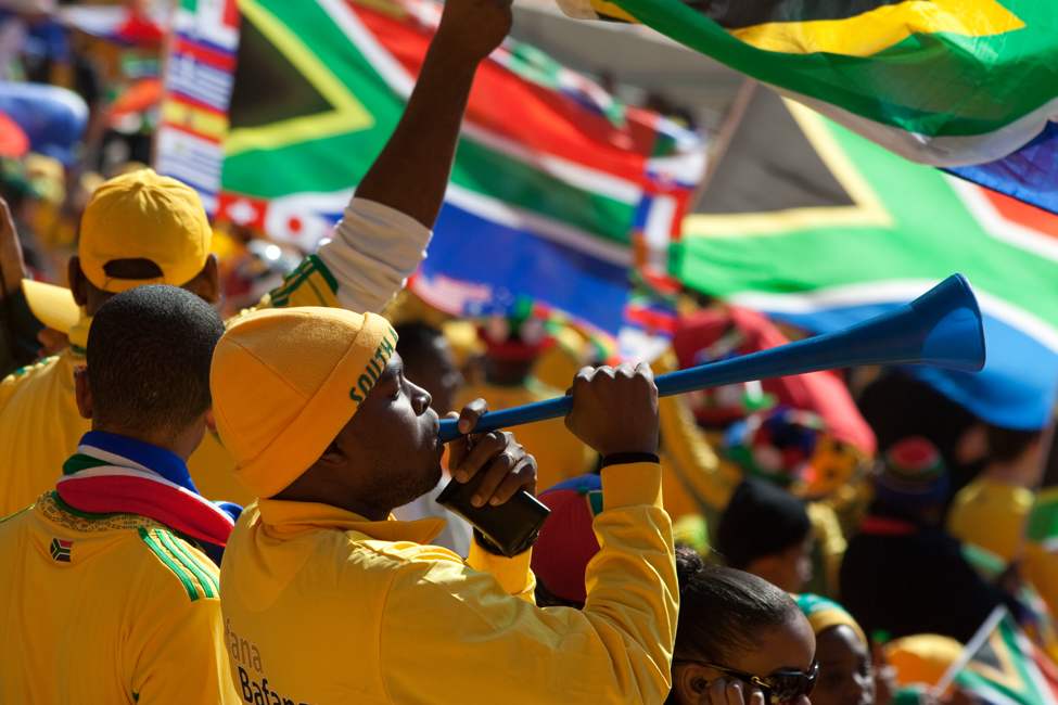 spectator in stadium blowing long plastic horn as South African national flags flying in the background