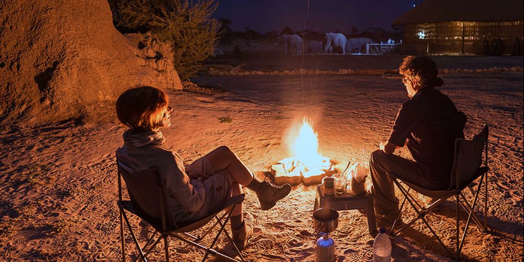 Two people sit in camp chairs next to a small campfire at night with elephants visible in the distance