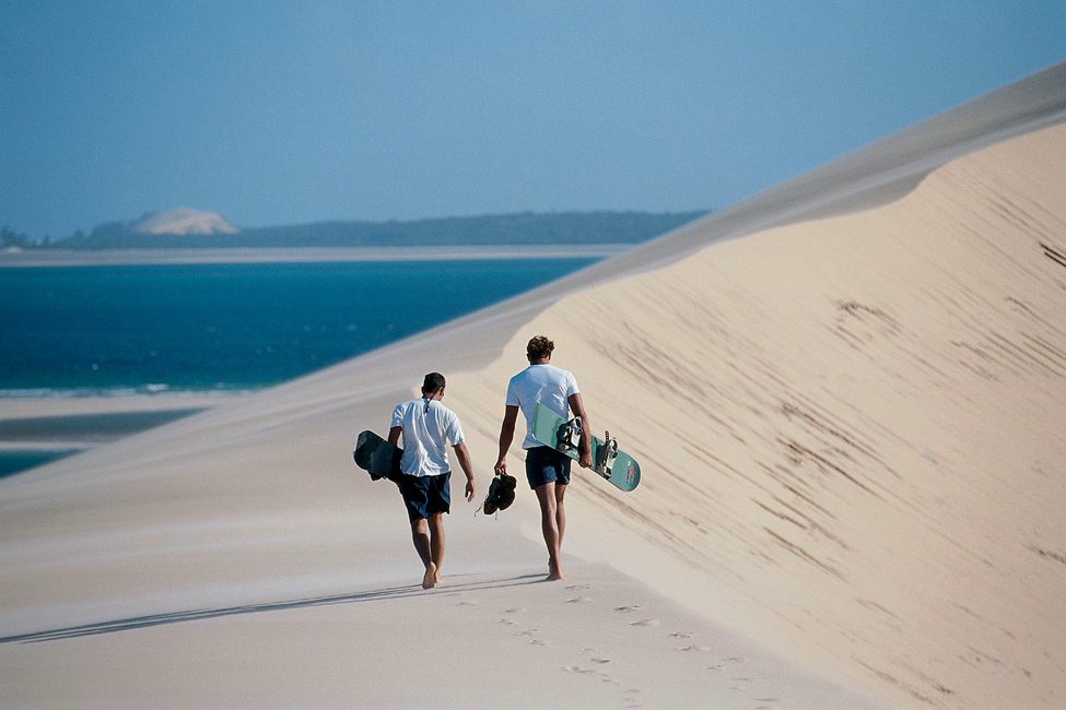 two individuals carrying dune boards on a sand dune near the ocean on Bazaruto Islans