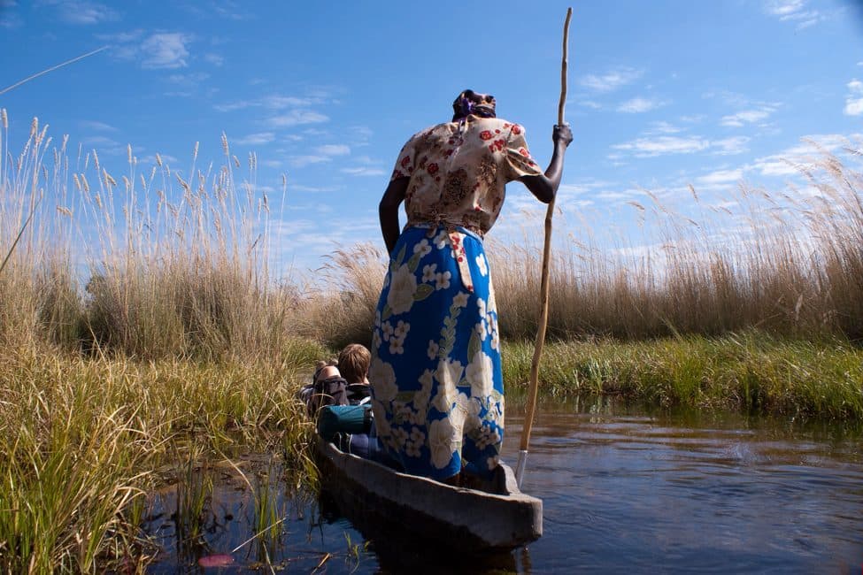 Person in red flowered shirt and blue flowered skirt steers a dugout canoe through the Okavango Delta