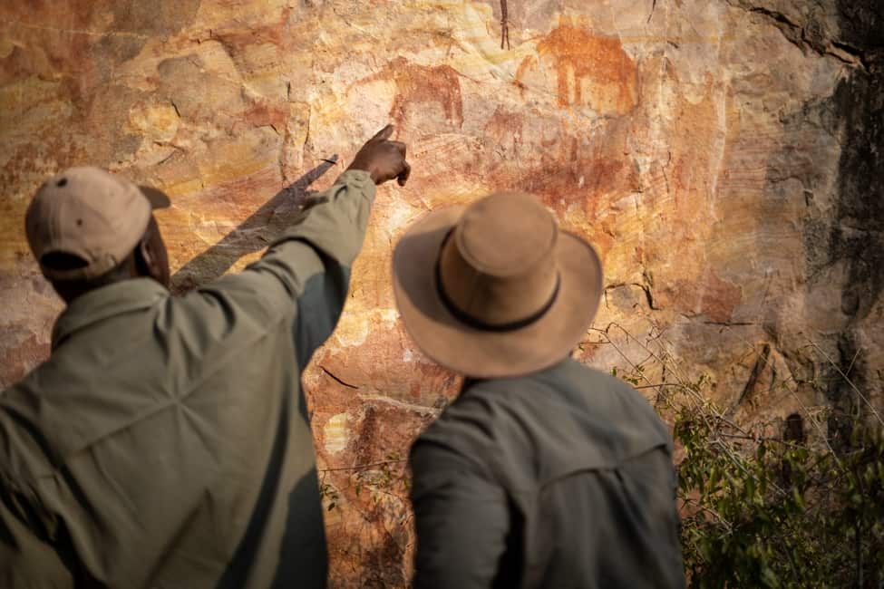 Safari guide points to rock paintings of elephant silhouettes. The paintings are in red and orange ochre-type colors on sandstone background.