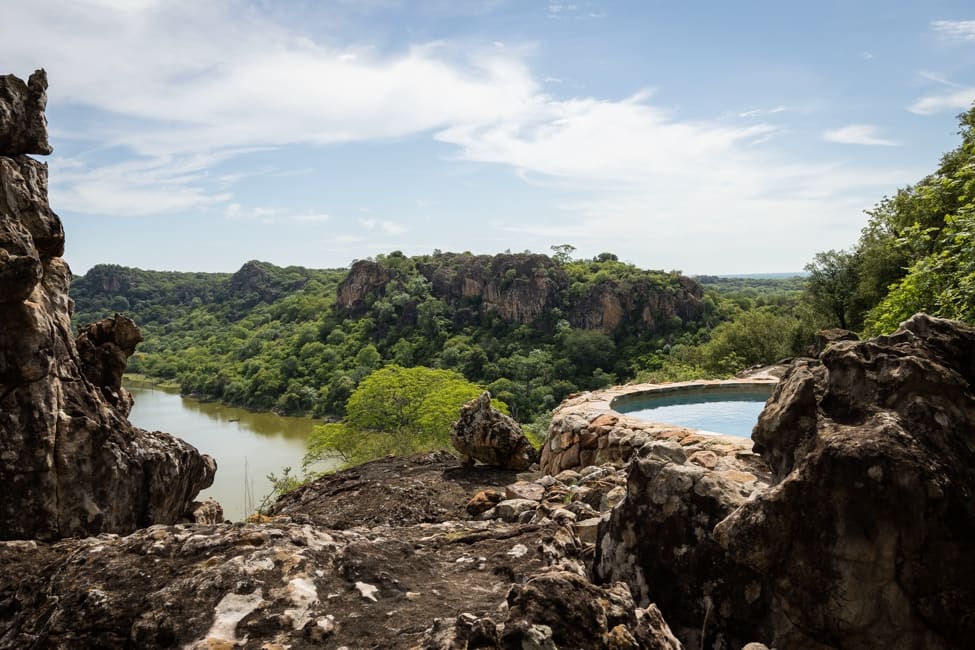 a round, stone-rimmed swimming pool built on top of a hill overlooks a river and green, tree-covered valley